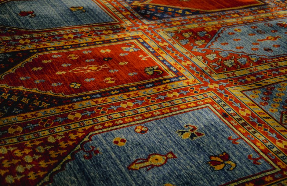 Close-up view of a traditional oriental rug with intricate patterns in shades of red, orange, blue, and yellow, laid out on a flat surface. The carpet's detailed floral and geometric motifs are clearl