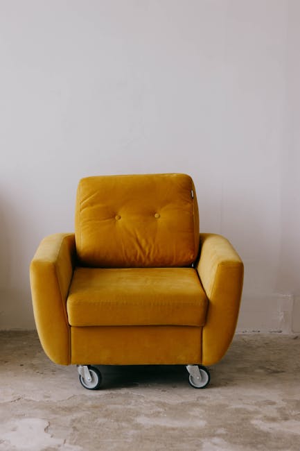 A mustard yellow armchair with a vintage design, featuring a cushioned backrest with button tufting, situated on a concrete floor against a plain white wall. The chair has rounded armrests and is moun